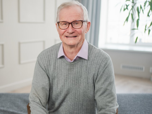 Smiling older man with white hair and glasses, wearing a light gray sweater and pastel purple shirt, indoors.